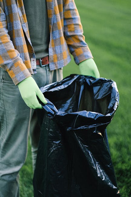 A person standing outdoors on a grassy area, wearing a plaid shirt over a gray t-shirt, light-colored trousers, and bright green gloves, is holding open a large black trash bag lined with a shiny plastic interior. The individual appears to be preparing to dispose of rubbish, with the bag partially expanded and empty. The scene suggests an activity related to rubbish removal or waste collection, highlighting the practical use of the bag for rubbish disposal in an outdoor setting. The background features a well-maintained, lush green lawn, with natural daylight illuminating the scene, emphasizing the context of waste management or outdoor cleaning services possibly provided by a company like Rubbish Removal Balham.