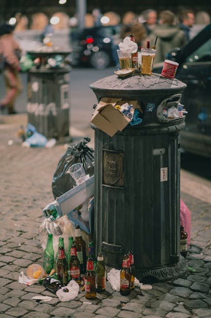 A black cylindrical public rubbish bin made of metal with vertical ridges and a domed top sits on a cobblestone street, surrounded by various discarded waste. On top of the bin are used paper cups with lids and straws, a crumpled paper napkin, and a small cardboard container, indicating recent waste deposition. Attached to the side of the bin are a few clear plastic bags containing trash, and some cardboard pieces. At the base of the bin, there are several empty glass bottles, some of which are tipped over, along with crumpled paper and other litter scattered on the cobblestones. The background shows a busy street scene with blurred pedestrians, parked cars, and a larger trash container in the distance, suggesting an outdoor urban environment during daytime. The overall scene highlights typical roadside litter that can accumulate if not regularly collected, exemplifying the need for effective waste management services such as those offered by Rubbish Removal Balham, specializing in scheduled rubbish removal and private disposal solutions.