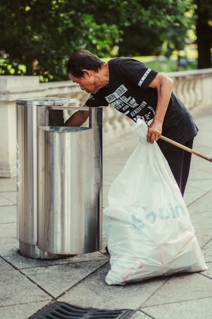 A person standing outdoors on a grassy area, wearing a plaid shirt over a gray t-shirt, light-colored trousers, and bright green gloves, is holding open a large black trash bag lined with a shiny plastic interior. The individual appears to be preparing to dispose of rubbish, with the bag partially expanded and empty. The scene suggests an activity related to rubbish removal or waste collection, highlighting the practical use of the bag for rubbish disposal in an outdoor setting. The background features a well-maintained, lush green lawn, with natural daylight illuminating the scene, emphasizing the context of waste management or outdoor cleaning services possibly provided by a company like Rubbish Removal Balham.