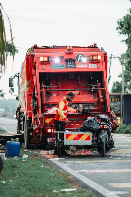 A worker in a red vest with yellow sleeves is standing at the rear of a large red rubbish collection truck, which is parked adjacent to a paved street. The truck features a metal cylindrical body with hydraulic lifting mechanisms and safety lights on top, and appears to be designed for waste collection and disposal. The worker is leaning slightly forward, handling a black trash bag or collection container positioned on a small wheeled platform nearby. The surrounding environment includes a section of road with a curb, some scattered waste on the ground, and a bicycle parked next to the worker. In the background, there are trees, utility poles with overhead wires, and distant buildings, suggesting an urban or suburban setting. The scene is illuminated by natural daylight, creating soft shadows and highlighting the textures of the materials, including the glossy finish of the truck, the fabric of the worker's vest, and the plastic of the waste containers. This image illustrates an independent rubbish collection operation involving a waste management worker, a collection vehicle, and necessary equipment, aligning with private waste handling services.