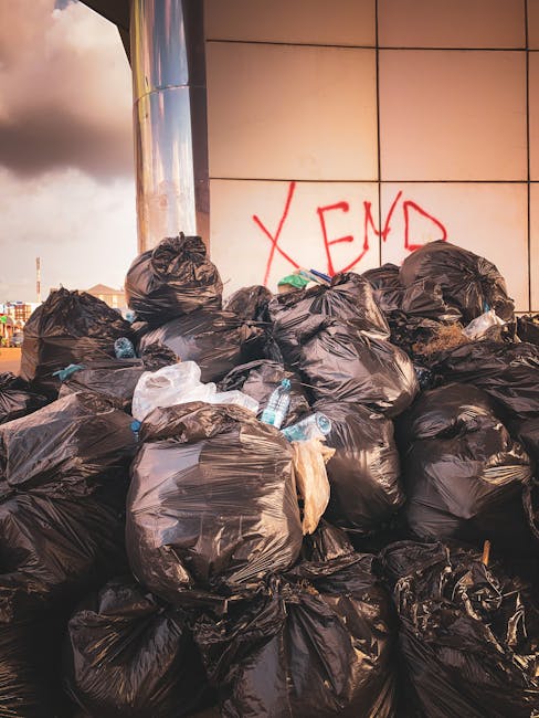 A large pile of black plastic rubbish bags stacked against a tiled wall outdoors, with some bottles and plastic waste visible on top and around the bags. The bags appear to be filled with household or general waste, and the tiles behind are a mix of beige and light brown, with a red spray-painted message that partially reads 'XEND' in the background. The scene is illuminated with natural light, highlighting the texture of the tightly tied bags and the glossy finish of the plastic. This setting suggests an instance of private waste disposal or waste collection from an outdoor area, possibly as part of rubbish removal services like those provided by Rubbish Removal Balham. The environment appears urban, with the waste positioned on a pavement or driveway adjacent to a modern building, emphasizing the need for effective rubbish removal or alternative waste handling solutions.