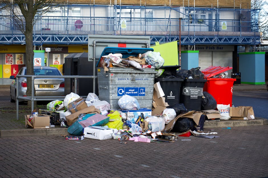A worker in a red vest with yellow sleeves is standing at the rear of a large red rubbish collection truck, which is parked adjacent to a paved street. The truck features a metal cylindrical body with hydraulic lifting mechanisms and safety lights on top, and appears to be designed for waste collection and disposal. The worker is leaning slightly forward, handling a black trash bag or collection container positioned on a small wheeled platform nearby. The surrounding environment includes a section of road with a curb, some scattered waste on the ground, and a bicycle parked next to the worker. In the background, there are trees, utility poles with overhead wires, and distant buildings, suggesting an urban or suburban setting. The scene is illuminated by natural daylight, creating soft shadows and highlighting the textures of the materials, including the glossy finish of the truck, the fabric of the worker's vest, and the plastic of the waste containers. This image illustrates an independent rubbish collection operation involving a waste management worker, a collection vehicle, and necessary equipment, aligning with private waste handling services.