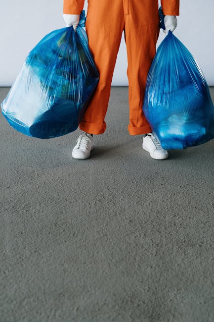 A person wearing orange work overalls and white sneakers is standing on a grey carpeted floor, holding two large blue plastic rubbish bags, one in each hand. The bags are filled with waste materials, visible through the semi-transparent plastic, and are slightly bulging at the top. The person's torso and head are not visible in the image. This scene appears to take place indoors, possibly in a storage area or warehouse. The setting suggests a private waste handling or rubbish collection activity, supported by the use of familiar domestic or commercial rubbish bags. The image conveys a focus on waste disposal or rubbish removal, consistent with services offered by companies like Rubbish Removal Balham, and highlights the practicality of manual collection for bulky or accumulated waste, aligning with the context of independent collection and alternative waste management options.