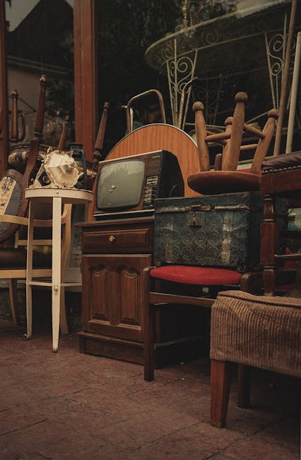 A collection of various old and spare furniture items arranged outdoors on a paved surface, including wooden chairs with different finishes and upholstery, a small vintage television with a rounded screen and dark casing, a blue patterned fabric storage box, a white side table with a skull ornament on top, and a wooden cabinet with paneled doors. The furniture appears to be set against a background of garden trees and a metal-framed circular patio table with intricate scrollwork detailing. The scene suggests a furniture clearance or private collection for disposal, typical of rubbish removal services like those provided by Rubbish Removal Balham. The natural lighting indicates daytime, with ambient light casting soft shadows over the items, emphasizing their textures and colors. This setup reflects a typical scenario of clearing unwanted household furnishings before disposal or professional collection, aligned with independent waste handling practices for bulky items outside of municipal collections.