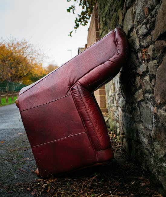 A large, three-seater leather sofa with a deep burgundy color and smooth, shiny surface sits upright against a rough, irregular stone wall on a paved outdoor pathway. The sofa's armrests and cushions show slight creases and wear, indicating prior use. The setting appears to be an outdoor residential area during autumn, with a background featuring trees with orange and yellow foliage, and a wooden fence along the left side of the pathway. The lighting is natural, with soft daylight illuminating the scene, creating subtle reflections on the leather surface. The sofa's position suggests it has been deliberately placed for disposal or removal, consistent with the activities of a waste management service such as Rubbish Removal Balham, which provides private rubbish clearance solutions. Surrounding debris includes scattered leaves and small twigs on the ground, further emphasizing the outdoor environment. This scene exemplifies the type of furniture items that might be handled during an on-site furniture clearance or alternative waste collection process, aligning with the context of independent rubbish removal services.
