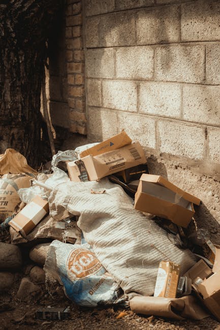 A pile of discarded rubbish placed against a brick and concrete wall outdoors, consisting of flattened cardboard boxes, some with visible print, and multiple white and beige plastic bags filled with waste. The cardboard boxes show folding edges and varying shades of brown, with some partially crushed or torn. The plastic bags appear bulky and are likely filled with additional rubbish, with some bags tightly knotted at the top. The scene includes a rough, uneven ground surface with small stones and dirt, and a tree trunk visible on the left side of the image. The wall behind the rubbish is constructed from concrete blocks with a light, slightly weathered finish, and neighboring bricks with a reddish-brown hue are partially visible on the left. The overall environment suggests an area designated for rubbish accumulation, possibly for collection or removal, aligning with the services offered by Rubbish Removal Balham for independent waste clearance or private disposal solutions.